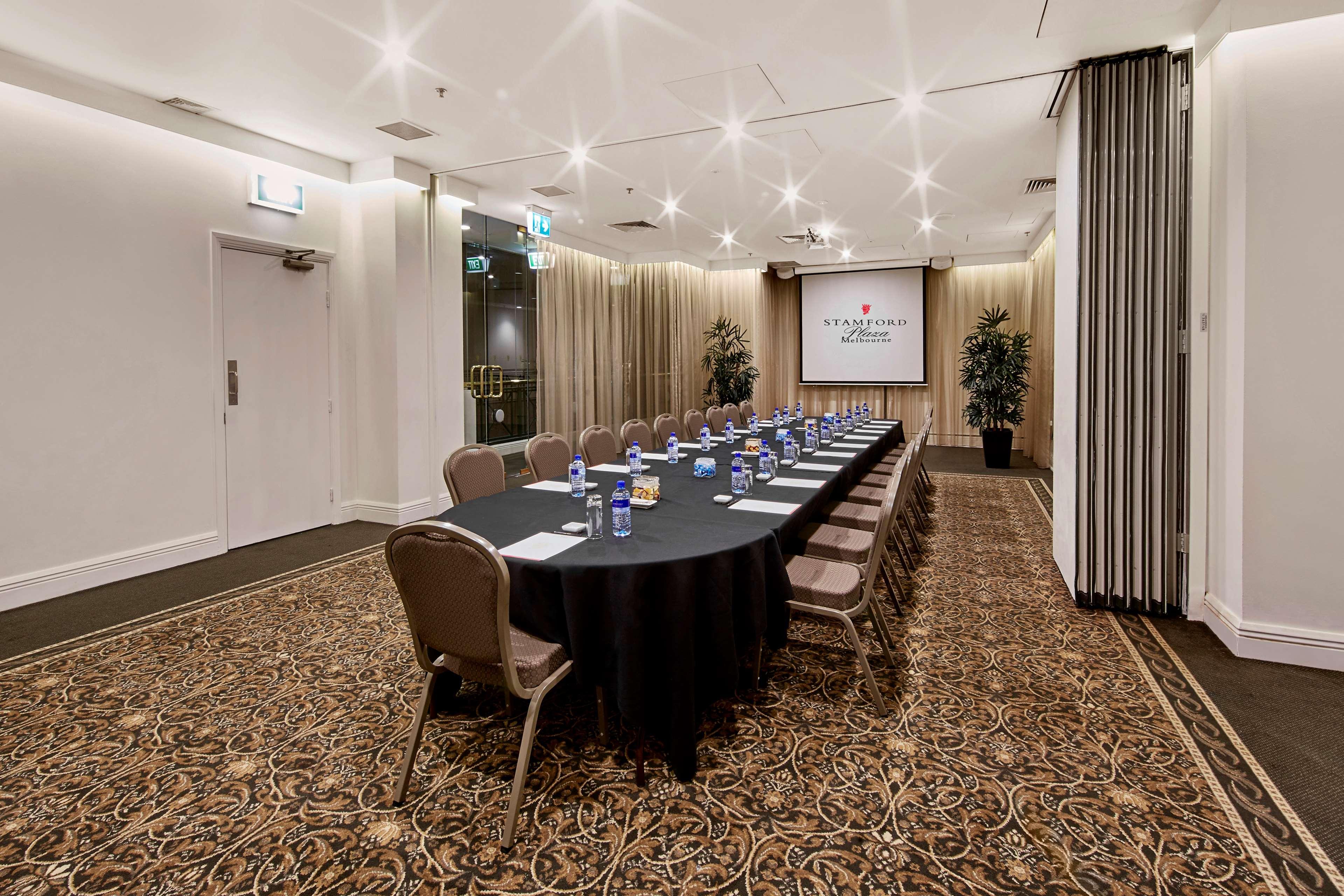 Stamford Plaza Melbourne Hotel Exterior foto The photo shows a meeting room set up for a conference or presentation. There is a long, rectangular table covered with a black tablecloth, surrounded by chairs. Each seat has a water bottle placed in front of it. At the front of the room, there is a